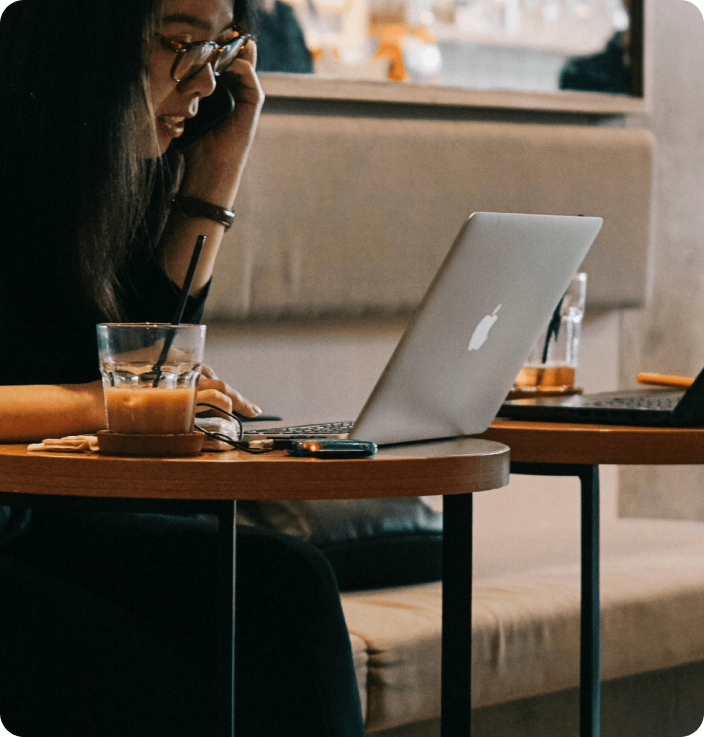 A young girl on the phone discussing work matters. There is a laptop and a cup of coffee on the table next to her.