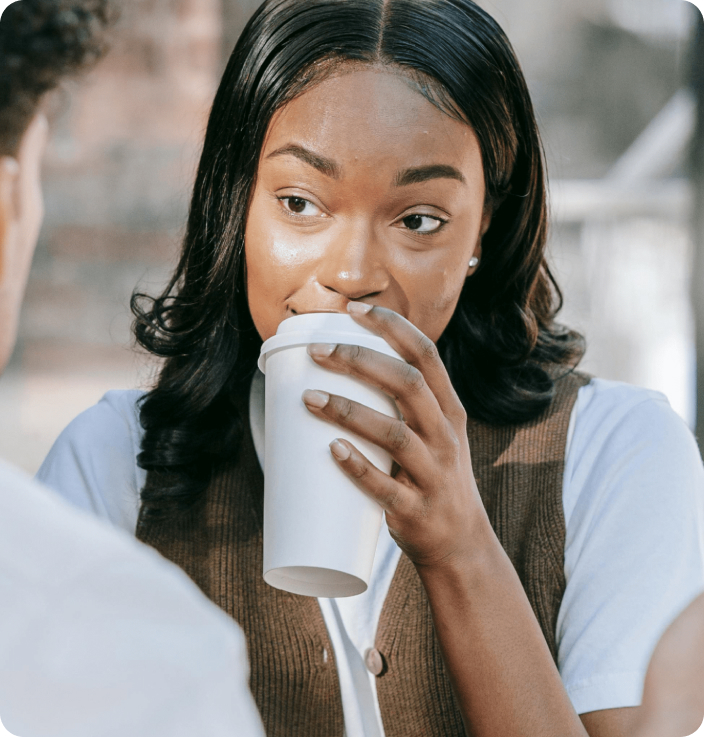 Woman in White Shirt with Brown Vest Drinking Coffee with a Man Seated in Front of Her